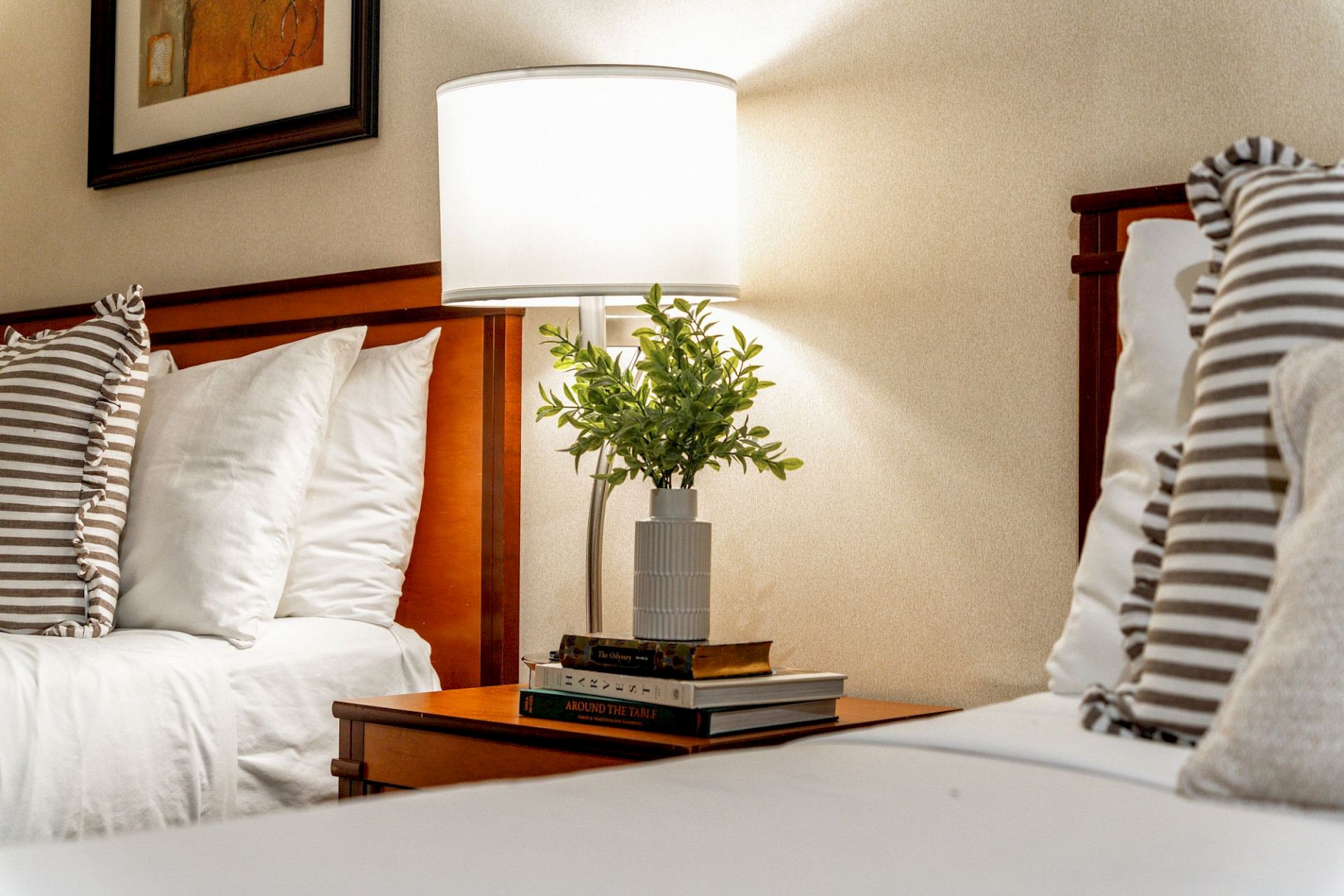 A hotel-style bedroom scene: a lit table lamp, a potted plant, stacked books, and striped pillows on a bed.