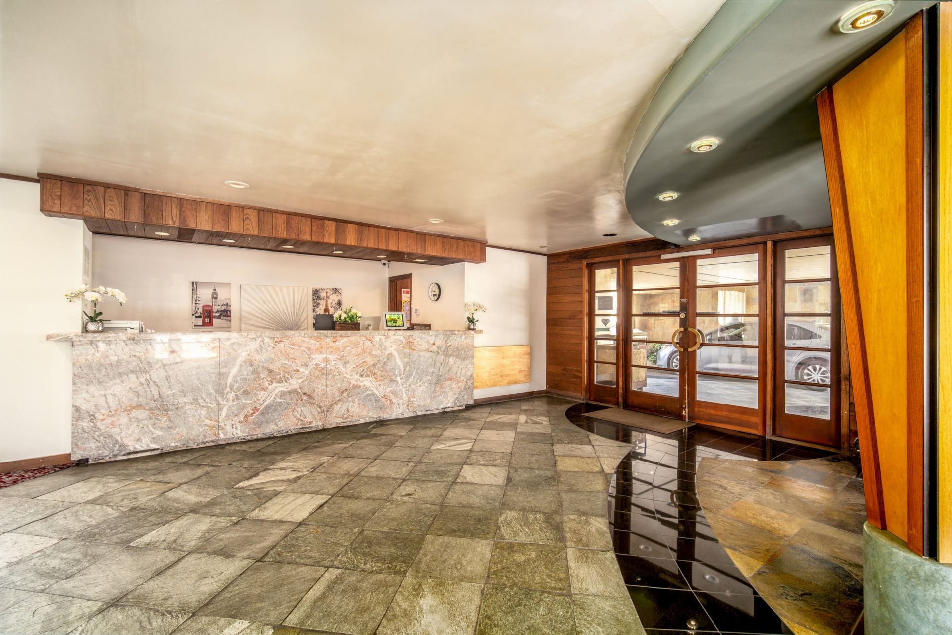 A hotel lobby with a marble reception counter, wood walls, polished stone floors, ceiling lights, and glass entrance doors.