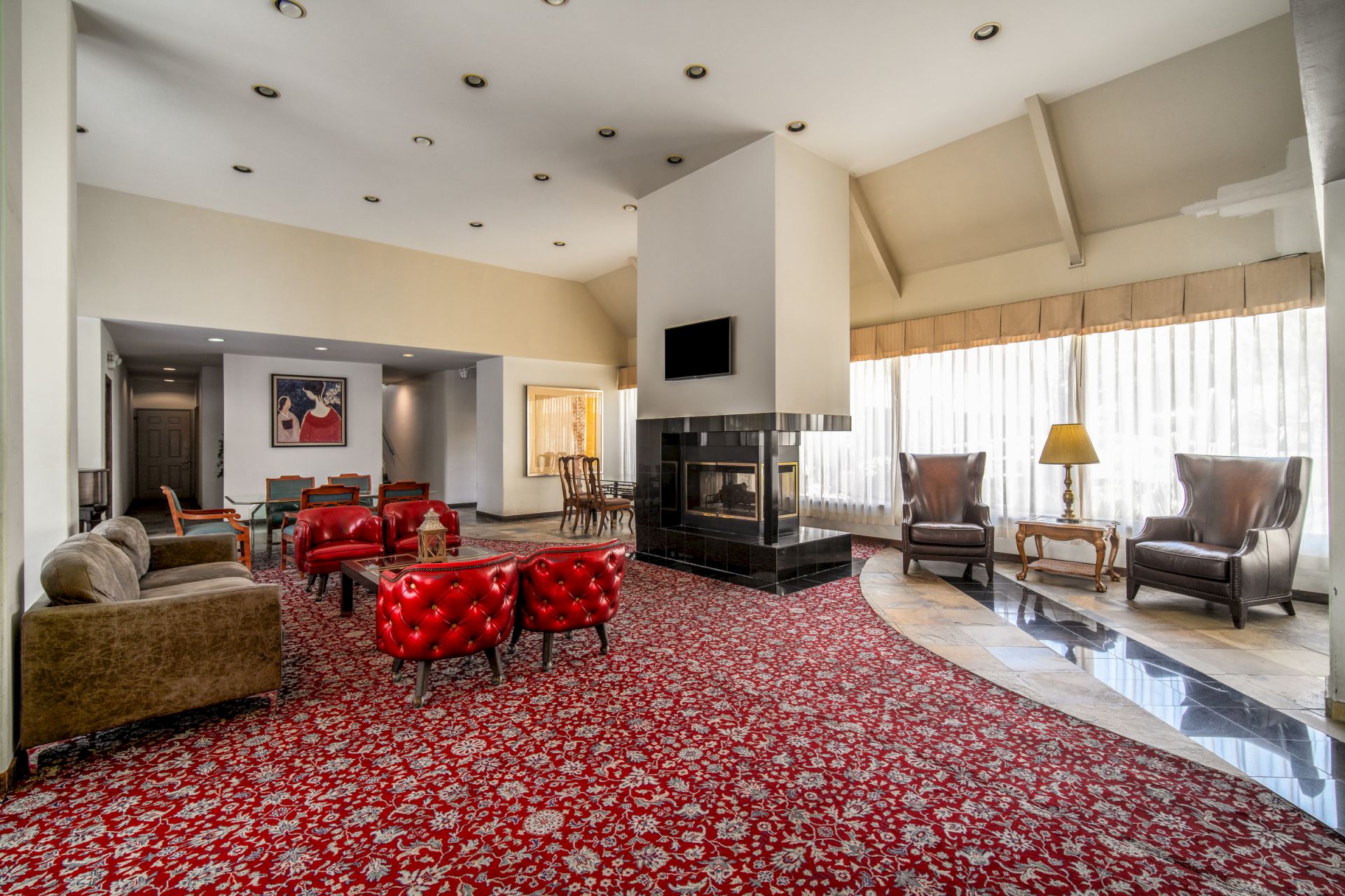 Bright living room with red carpet, leather seating, ornate fireplace, patterned rug, and floor lamps by large windows.