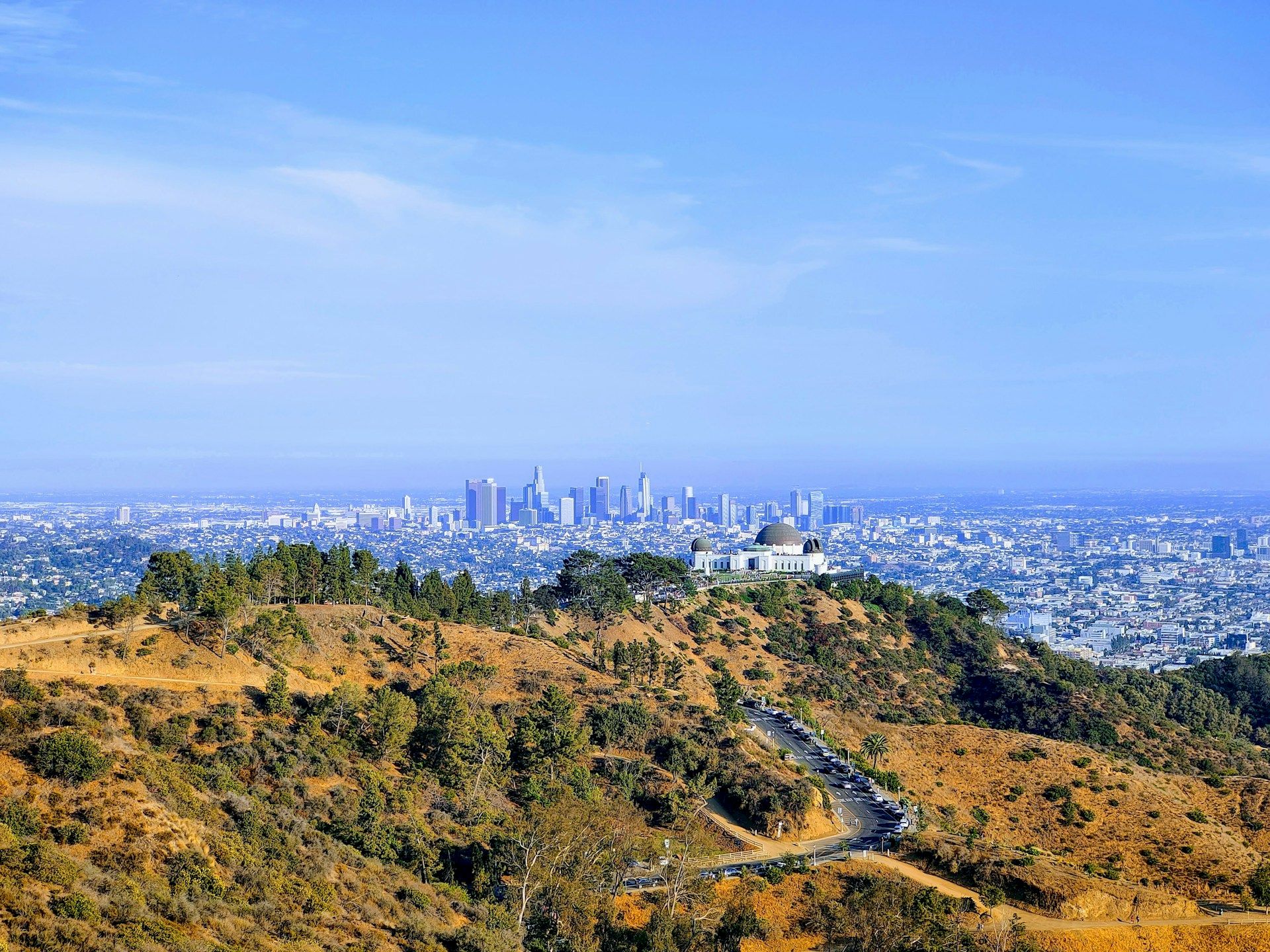 A panoramic view of a sprawling city skyline with a white observatory dome in the foreground hills beneath a clear blue sky.