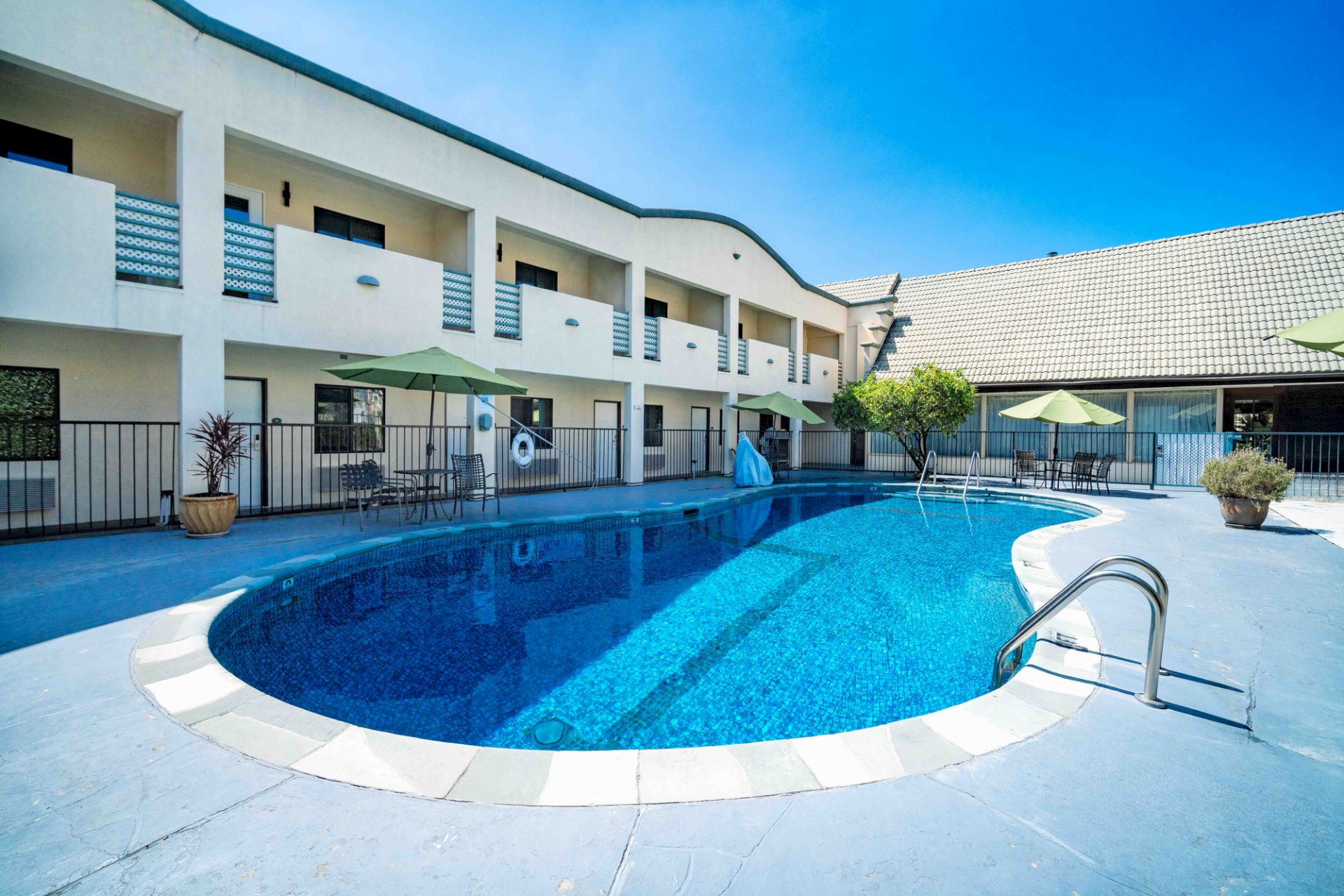 An outdoor courtyard pool with blue water, lounge chairs, green umbrellas, and white apartment buildings under a clear sky.
