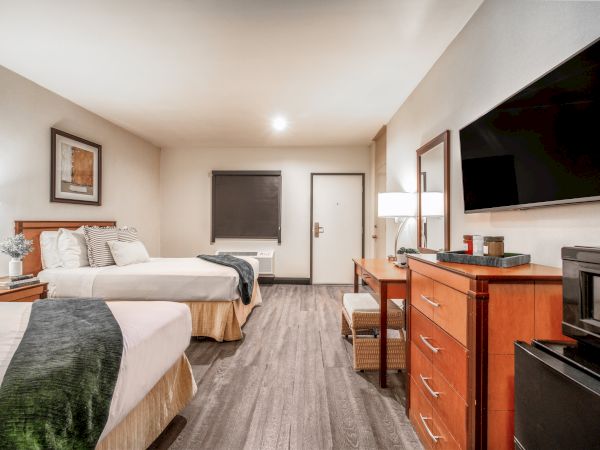 A bright hotel-style bedroom with a bed, nightstand, dresser, wall mirror, and a big flat-screen TV.