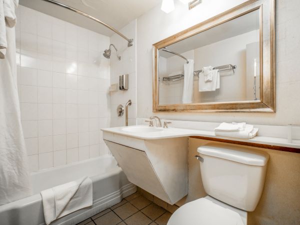 A bright hotel bathroom with a bathtub, showerhead, sink, towels, and a large gold-framed mirror.