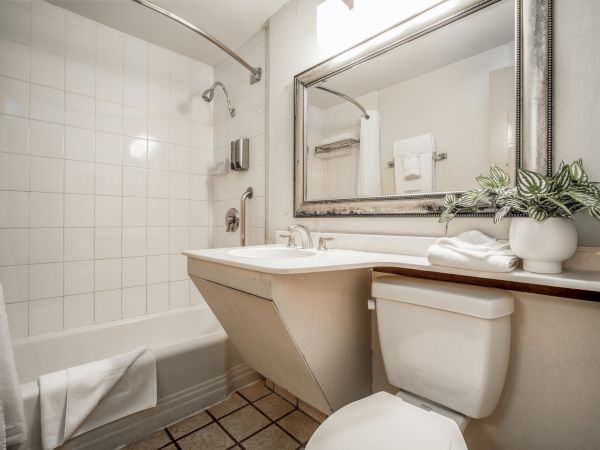 Bright bathroom with a bathtub, white tile walls, a sink, toiletries, and a large framed mirror above the toilet.