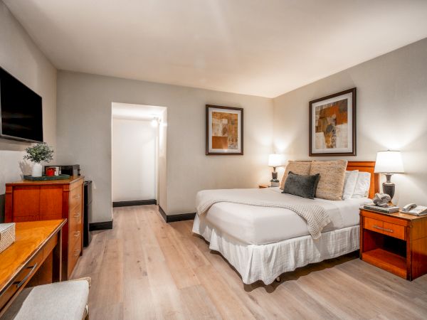 A bright hotel-style bedroom with a neatly made bed, bedside lamps, framed wall art, a TV cabinet, and light wood flooring.