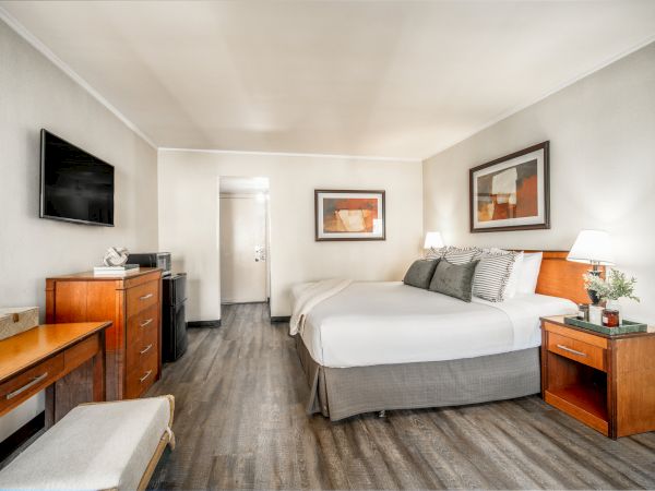 A tidy hotel-style bedroom with a king bed, wall-mounted TV, wooden dresser and bedside tables, lamps, and framed artwork.