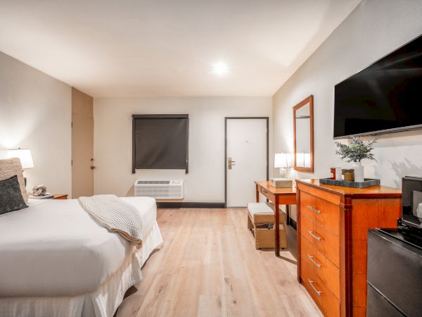 A bright hotel-style bedroom with a bed, wall TV, wooden dresser, mirror, and blackout shades over a window.