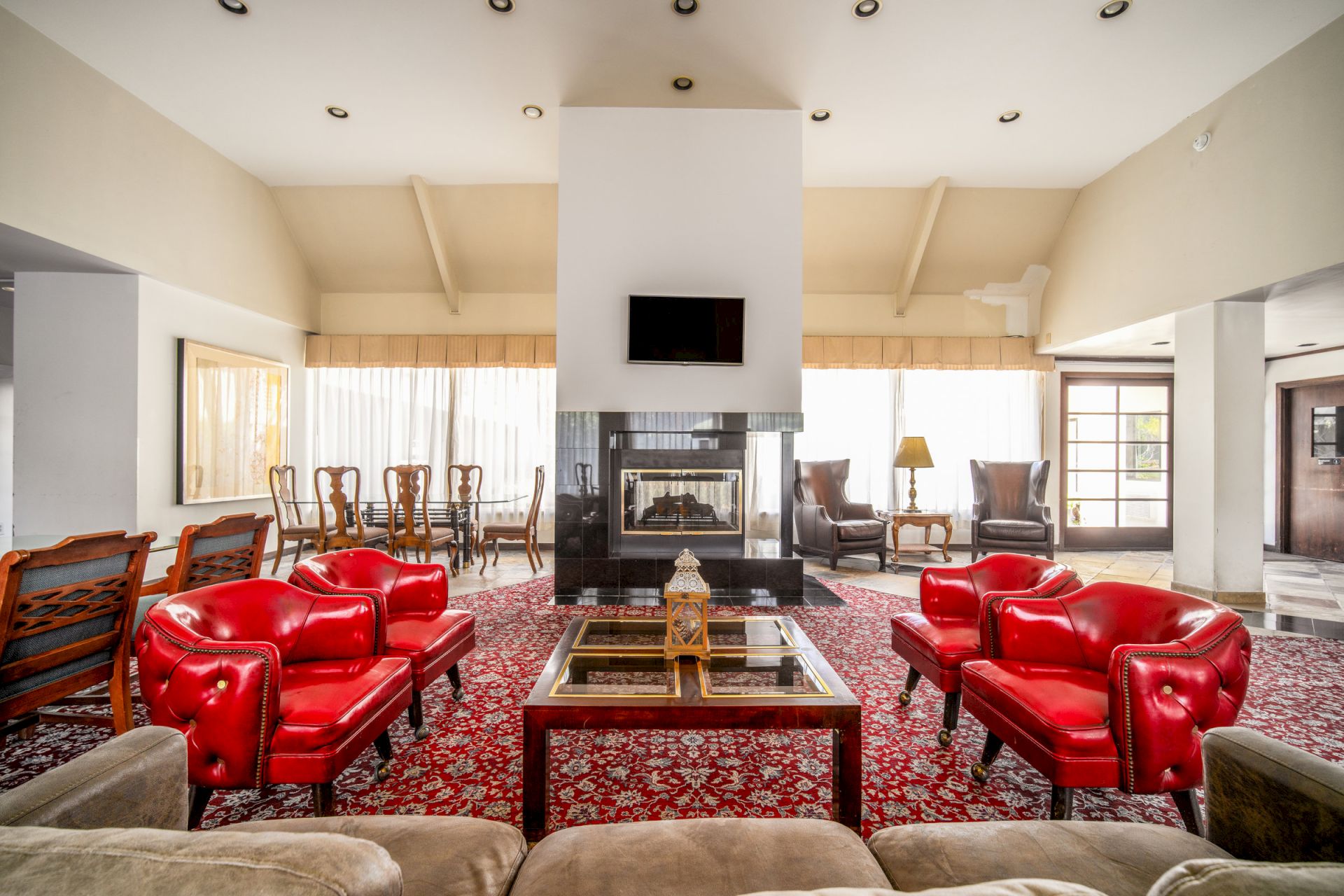A bright living room with a fireplace, flat-screen TV, red leather sofas, and a patterned rug with a central glass coffee table.