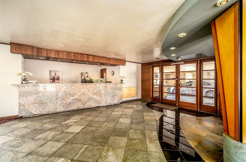 A hotel lobby with a marble reception counter, wood paneling, ceiling lights, and glass doors to the street outside.