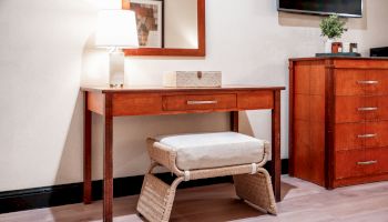 A cozy corner with a wooden desk, lamp, mirror, TV, side cabinet, plant, and a cushioned woven chair.