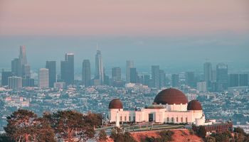 A sprawling city skyline at dusk with the Griffith Observatory domes in the foreground and tall skyscrapers behind.