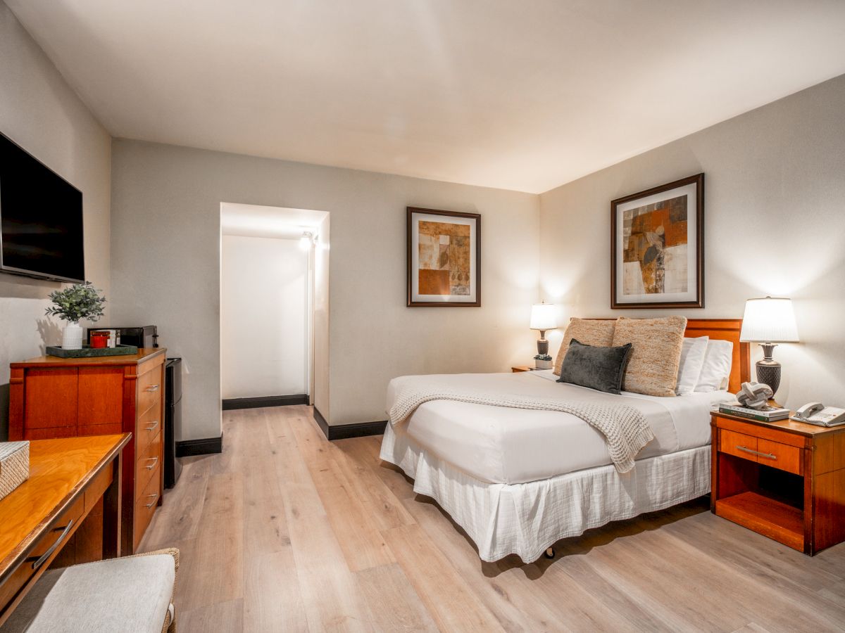 A bright hotel bedroom with a queen bed, nightstands, framed art, wall TV, and warm wood furniture.