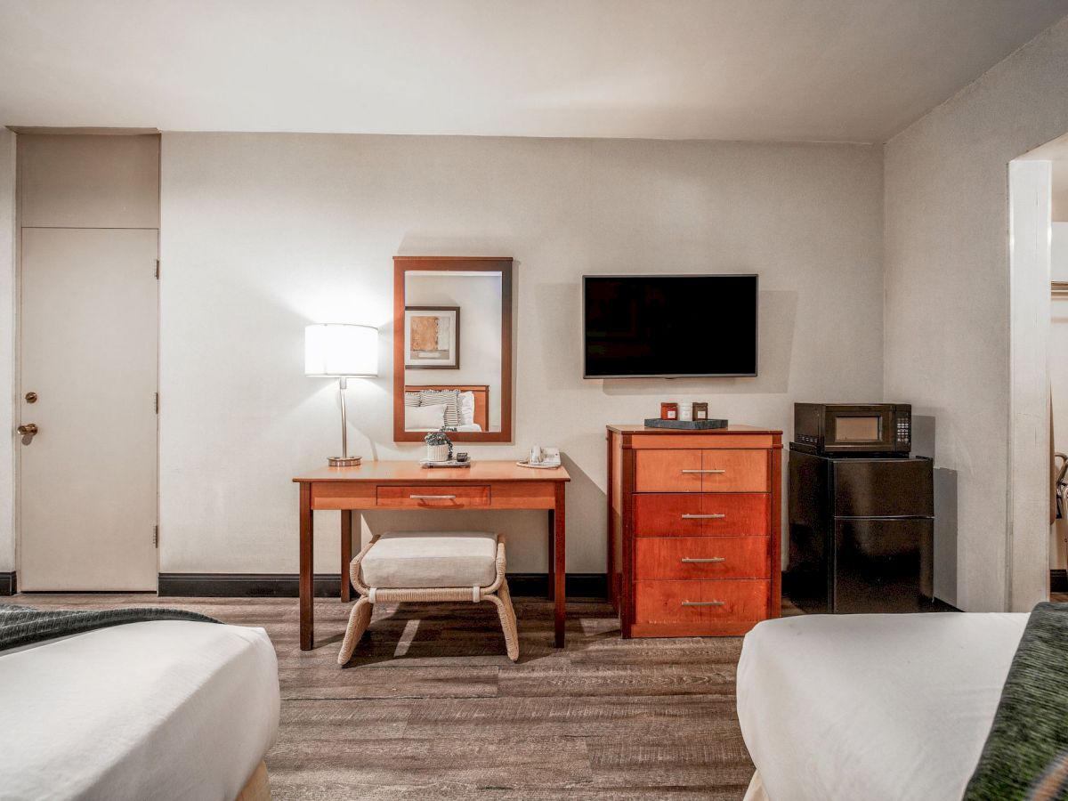 A tidy hotel room with two beds, a wooden desk, a lamp, a wall-mounted TV, and a cabinet holding coffee items.