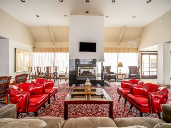 A bright luxury living room with a central fireplace, wall-mounted TV, red leather sofas, patterned rug, and warm wooden dining chairs.