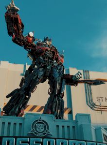 A towering red-and-blue sci‑fi robot (Transformers‑style) stands atop a building against a blue sky.