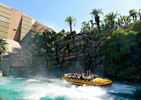 A crowd rides a yellow raft on a water-ride track, splashing past a tall slide wall in a tropical theme park.