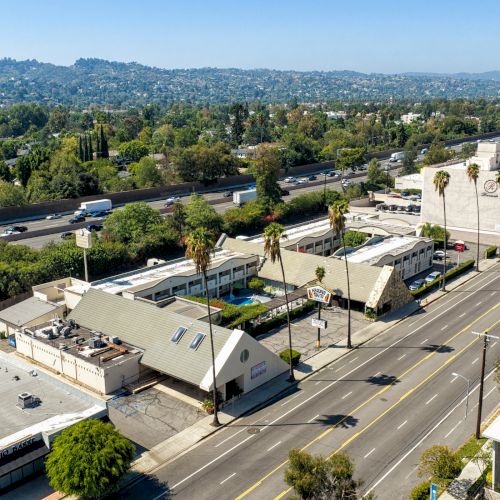 Aerial view of a busy highway with cars, low-rise buildings and greenery along rolling hills under a clear sky.