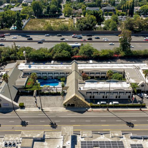 Aerial view of a hotel with a swimming pool and parking, beside a busy multi-lane highway and nearby homes.