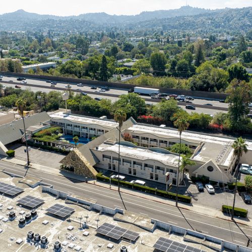 Aerial view of a busy highway with many cars beside industrial buildings and parking lots, set against green hills and trees.