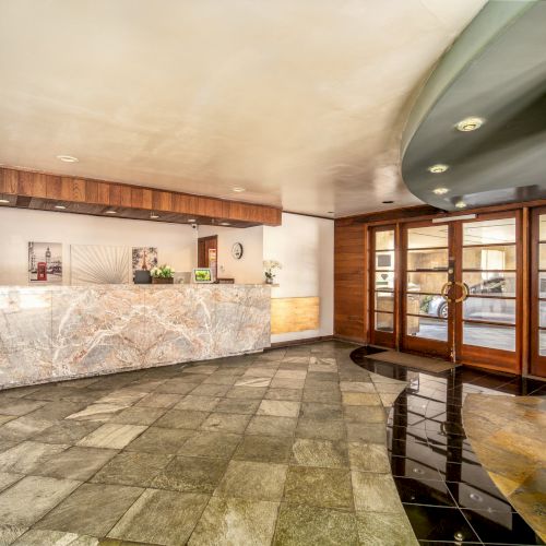 Indoor hotel lobby with a marble reception counter, wood paneling, warm lights, and a glass door to the street outside.