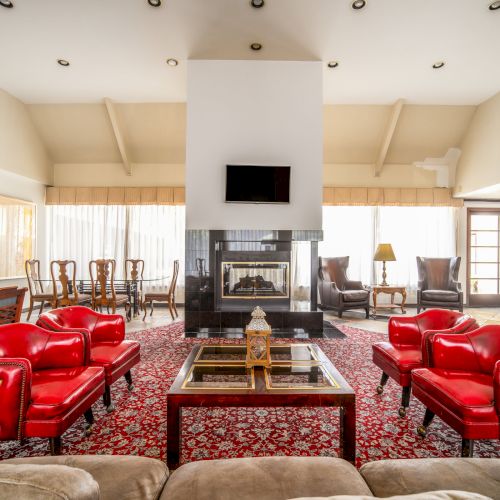 Bright living room with red leather sofas, a patterned rug, a central fireplace, and a mounted TV, plus chairs and curtains.