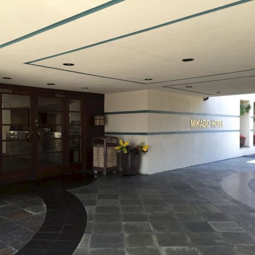 The entrance to the Mikado Hotel, with a tiled walkway, potted plants, dark glass doors, and a geometric ceiling.