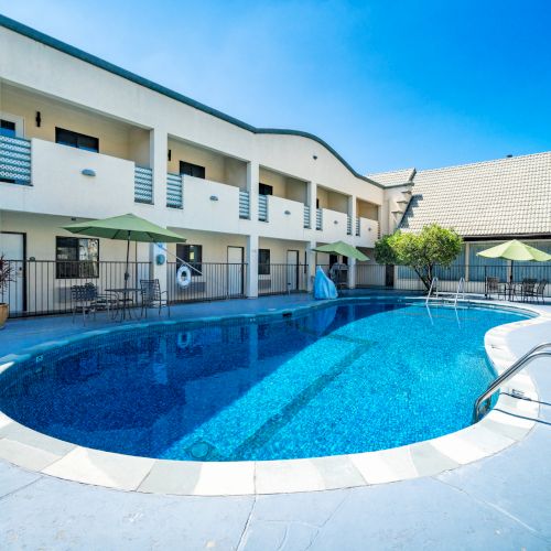 An outdoor swimming pool beside a modern building with green patio umbrellas and lounge chairs under a clear blue sky.