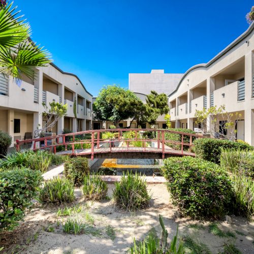 Sunny courtyard with palm trees, manicured greenery, a red bridge over a small tiled pool, and white apartment buildings under a blue sky.