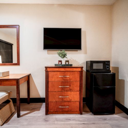 A hotel-style room corner with a flat-screen TV, wooden dresser, microwave/oven, fridge, table, lamp, and mirror, plus a small plant.