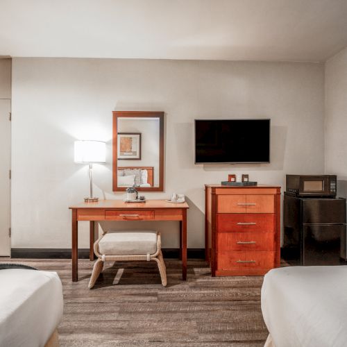 A hotel-style room with two beds, a wooden desk and lamp, a mounted flat-screen TV, and red cabinetry with a microwave/coffee station.