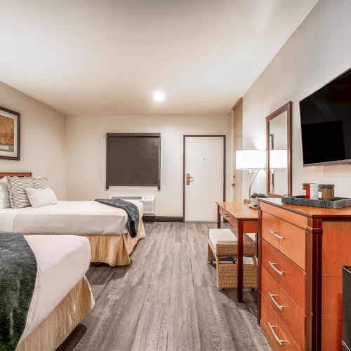 A bright hotel bedroom with a neatly made bed, dresser vanity, wall TV, and soft lighting in a modern suite.
