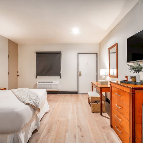 A bright hotel bedroom with a bed, dresser and flat-screen TV, plus a window, lamps, and a seating chair.