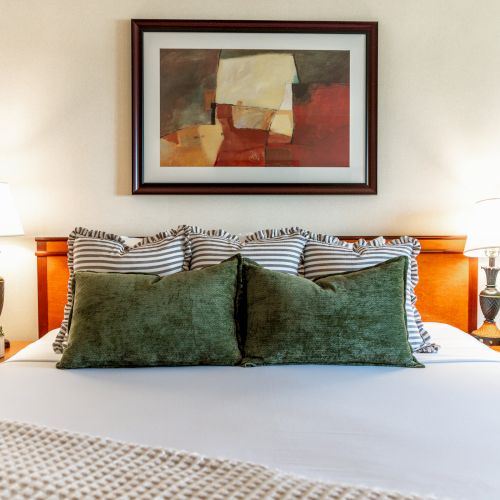 A cozy bedroom scene with a bed, green pillows, two lamps, and a framed abstract painting hanging above the headboard.