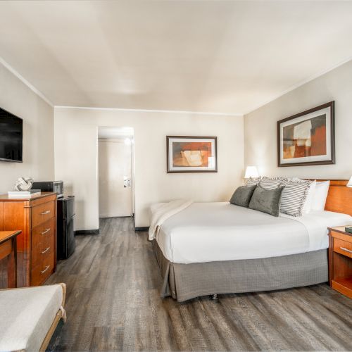 A tidy hotel bedroom with a queen bed, bedside tables, framed art, a wall TV, and a dresser in a modern neutral room.