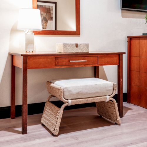 A wooden desk and dresser in a cozy home office, lit by a table lamp, with a woven chair and wall TV above.