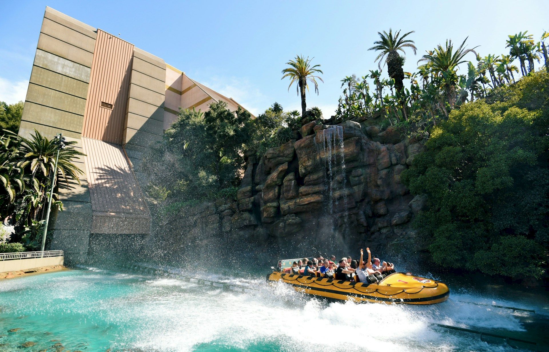 A group rides a yellow raft through a splashy water ride, with a waterfall backdrop, rocks, and palm trees.