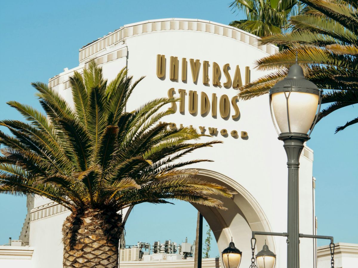 A Universal Studios sign reading &ldquo;Universal Studios&rdquo; with palm trees and a streetlamp under a clear blue sky.