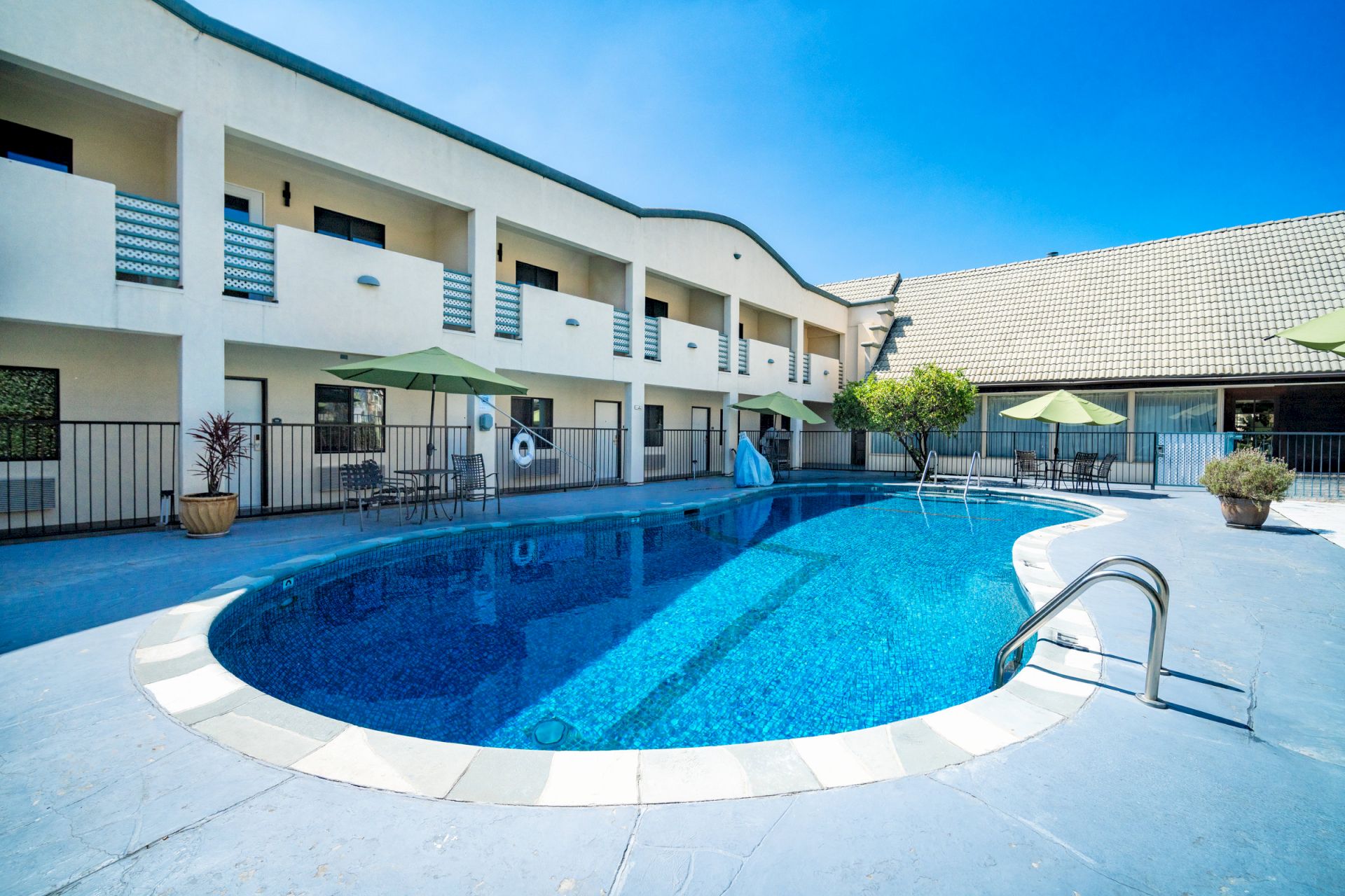 A blue tiled swimming pool beside a modern apartment building with green umbrellas and outdoor chairs.