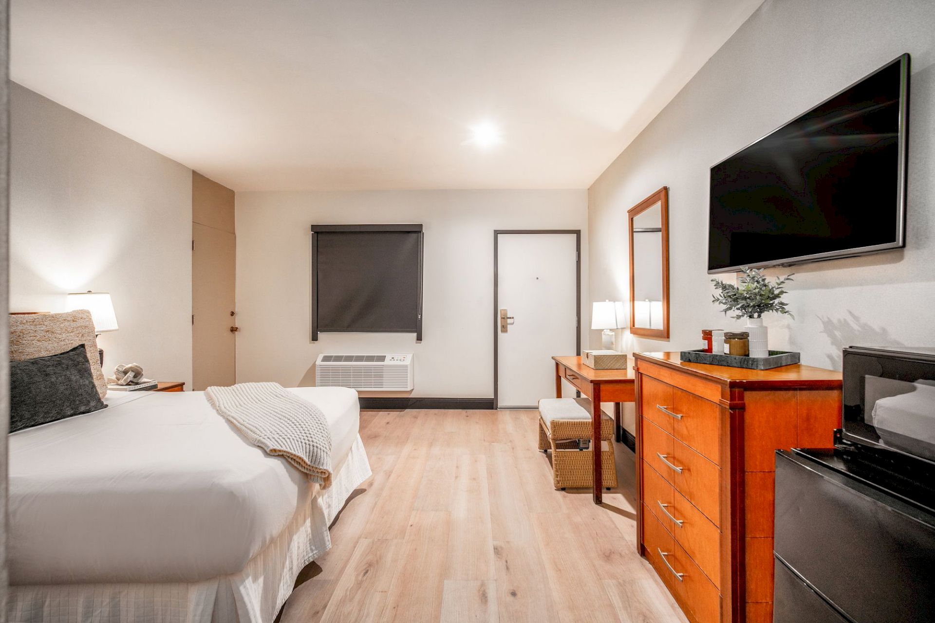 A bright hotel room with a bed, window, mini heater, TV, wooden dresser, and a vase on the counter.